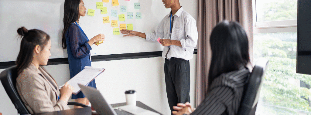 Designer leading a workshop at a whiteboard covered with sticky notes while teammates take notes, creative brief planning.