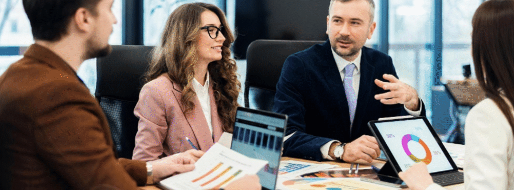 Marketing team discussing charts and print samples around a conference table, organizing a brand materials list.
